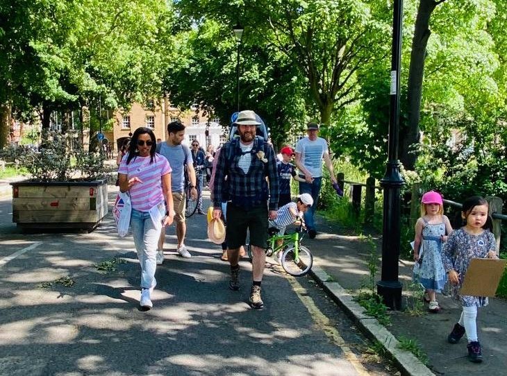 Adults and children walk under trees in a pedestrianised street on a sunny day.