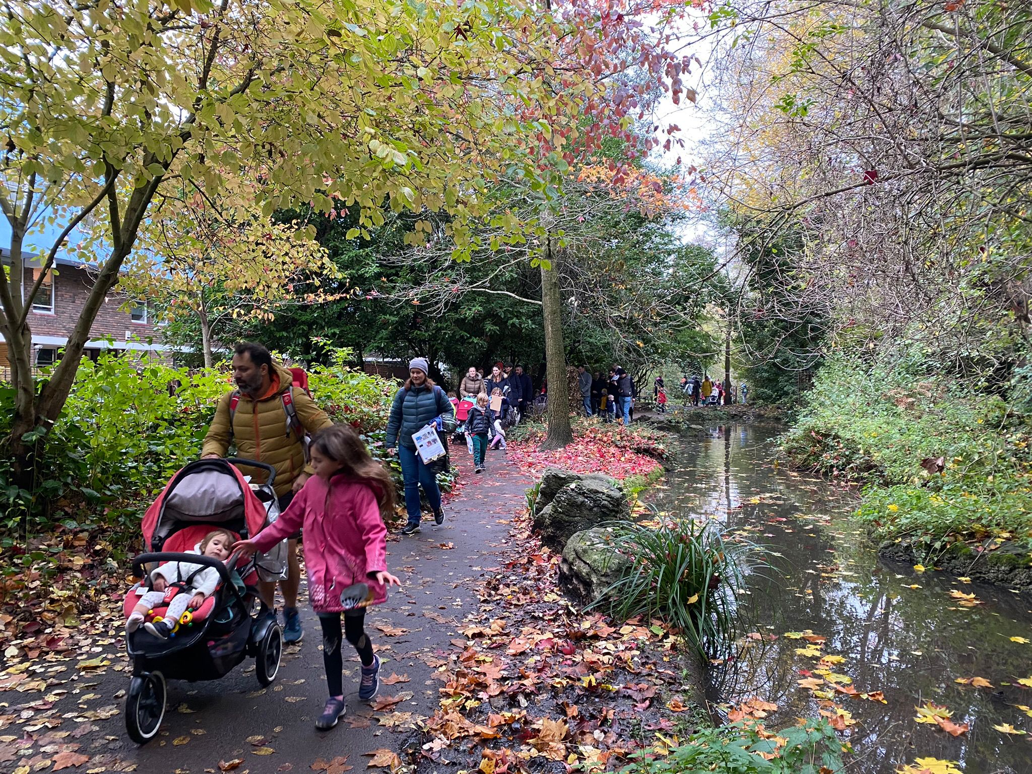 Families with children and prams walk along a leafy path next to a small, linear pond.