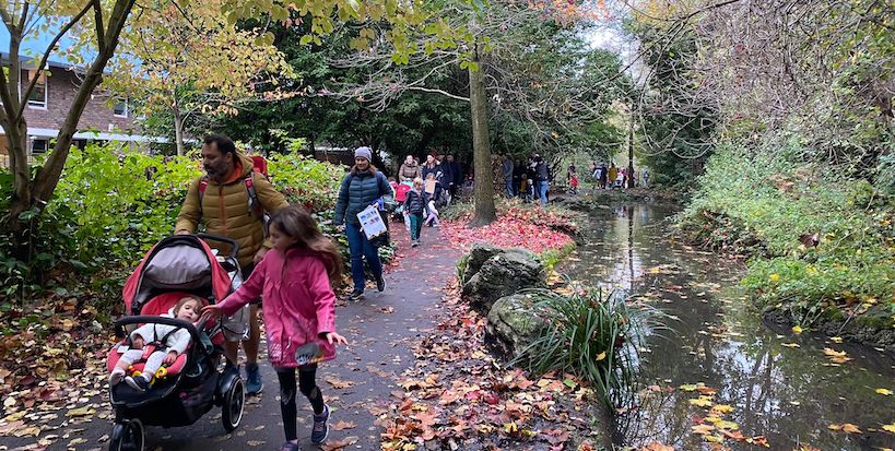 Families stroll along a linear pond with autumn leaves at their feet. There are trees and bushes all around.